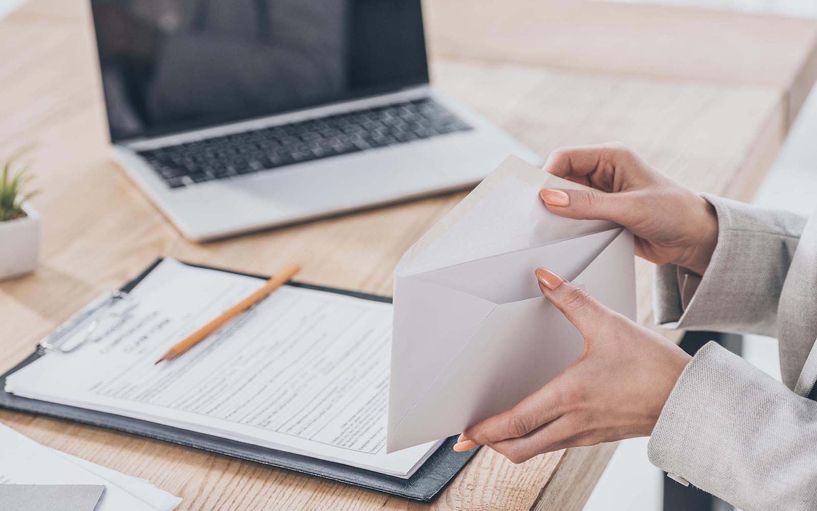 Person placing paper into an envelope while at a desk writing on a clipboard with laptop beside them