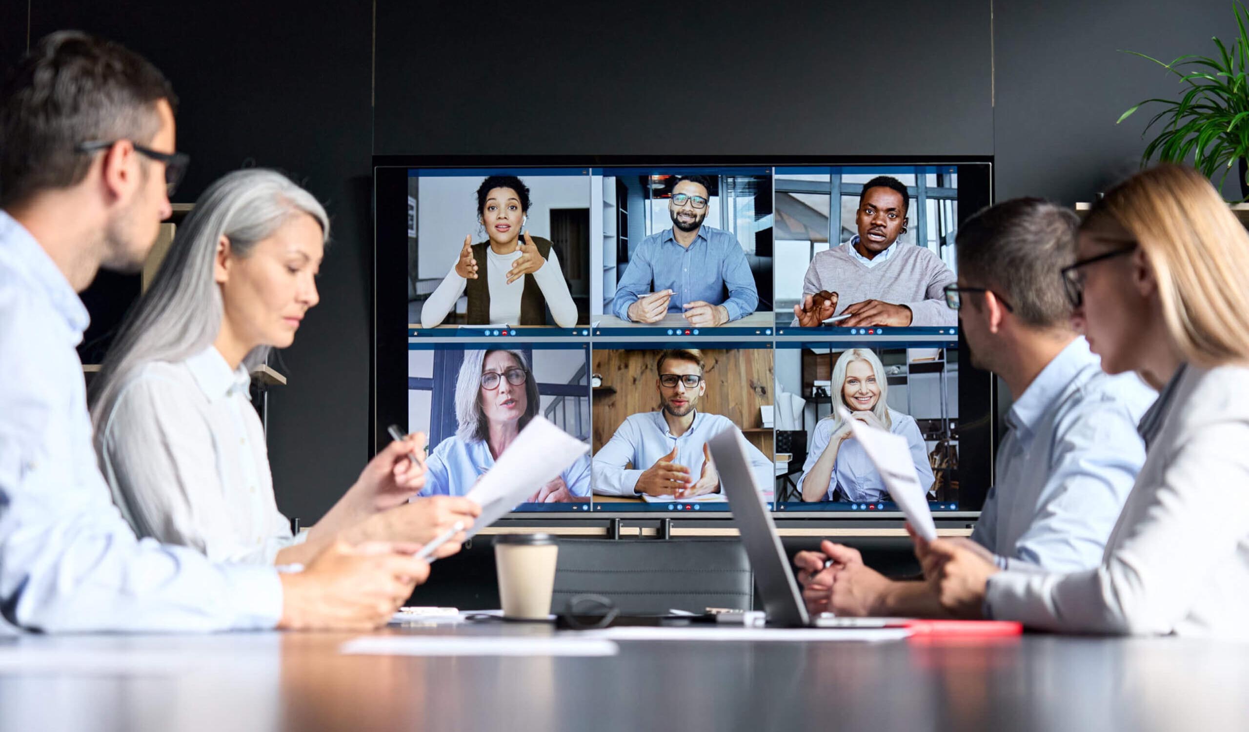 individuals having a hybrid meeting, half the team in person, half the team on a video call on a big screen in front of them